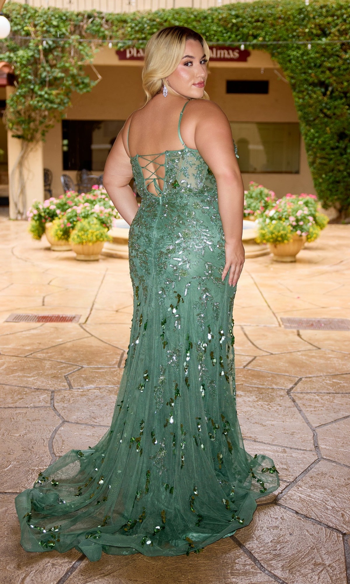 A woman in the Primavera 14106 Long Plus-Size Prom Dress, a green floor-length sequin gown with lace-up back detail, stands on a patio facing away from the camera.