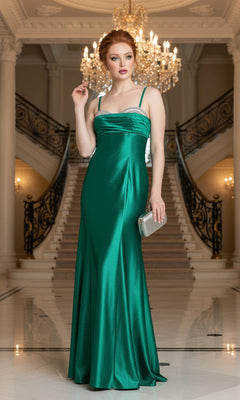Wearing the Morgan 14109 long emerald green prom dress with silver beaded details, a woman stands in a grand foyer beneath a chandelier, framed by dual staircases and holding a silver clutch.