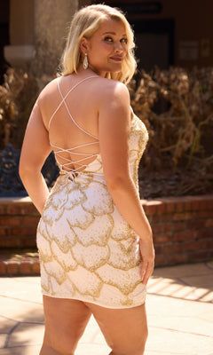 A woman poses outdoors in front of a brick planter with dry plants, wearing the Primavera 14115 Plus-Size Homecoming Dress—a short, light-colored mini dress featuring gold details, crystal beading, and a lace-up back.