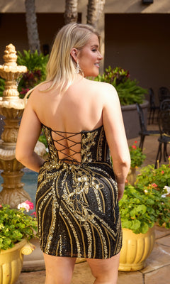 A woman stands outdoors surrounded by plants and a decorative fountain, wearing the Plus-Size Homecoming Dress: Primavera 14121—a black and gold sequin dress with a lace-up back.