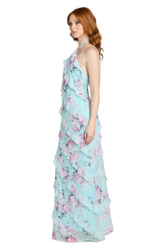 A woman stands in profile against a white background, wearing the Morgan 14173 Floral Ruffled Maxi Dress—a light blue, floor-length maxi featuring a pink floral print, ruffle tiers, halter neckline, and elegant open back that enhances its flowing silhouette.