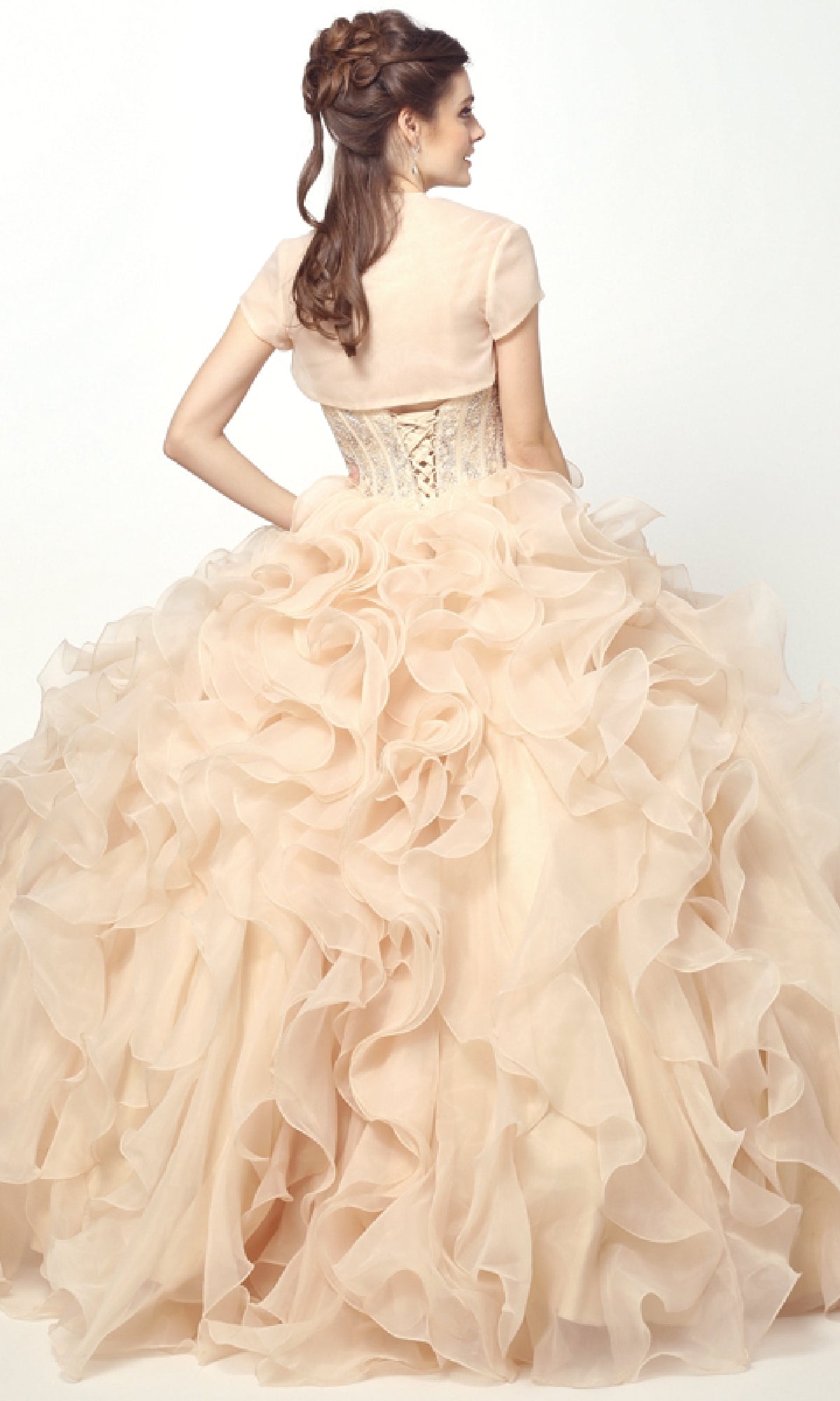 A woman stands facing away in the Champagne Quinceanera Dress 1418 By Juliet, featuring a light peach ruffled ball gown skirt and corset bodice. Her hair is styled in loose curls with a half-up braid, set against a plain white background.
