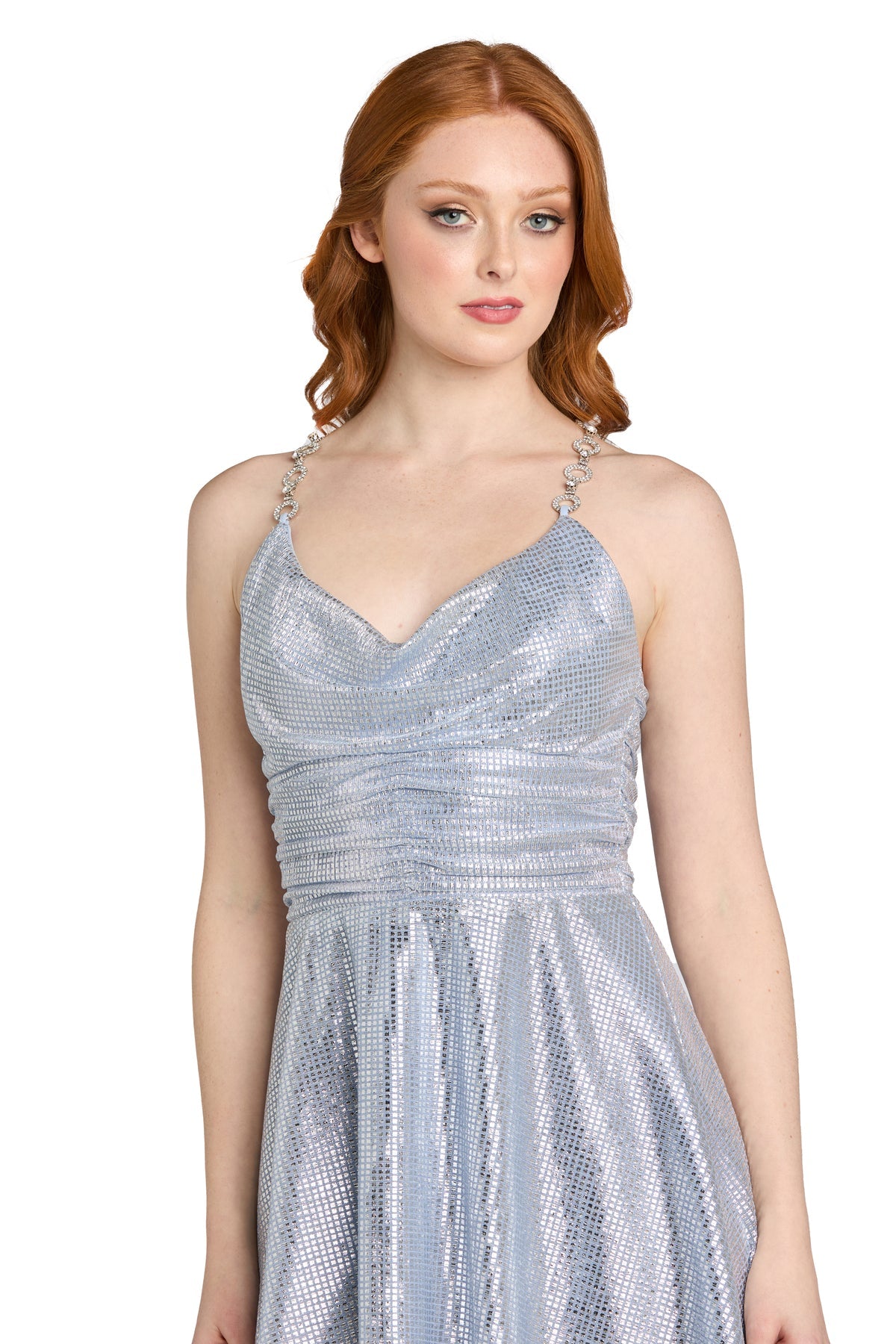 A woman with long red hair models the Morgan 14232 Short Metallic Homecoming Dress, featuring a silver sparkly fabric, decorative straps, and a chic cowl neckline, as she stands against a plain white background.