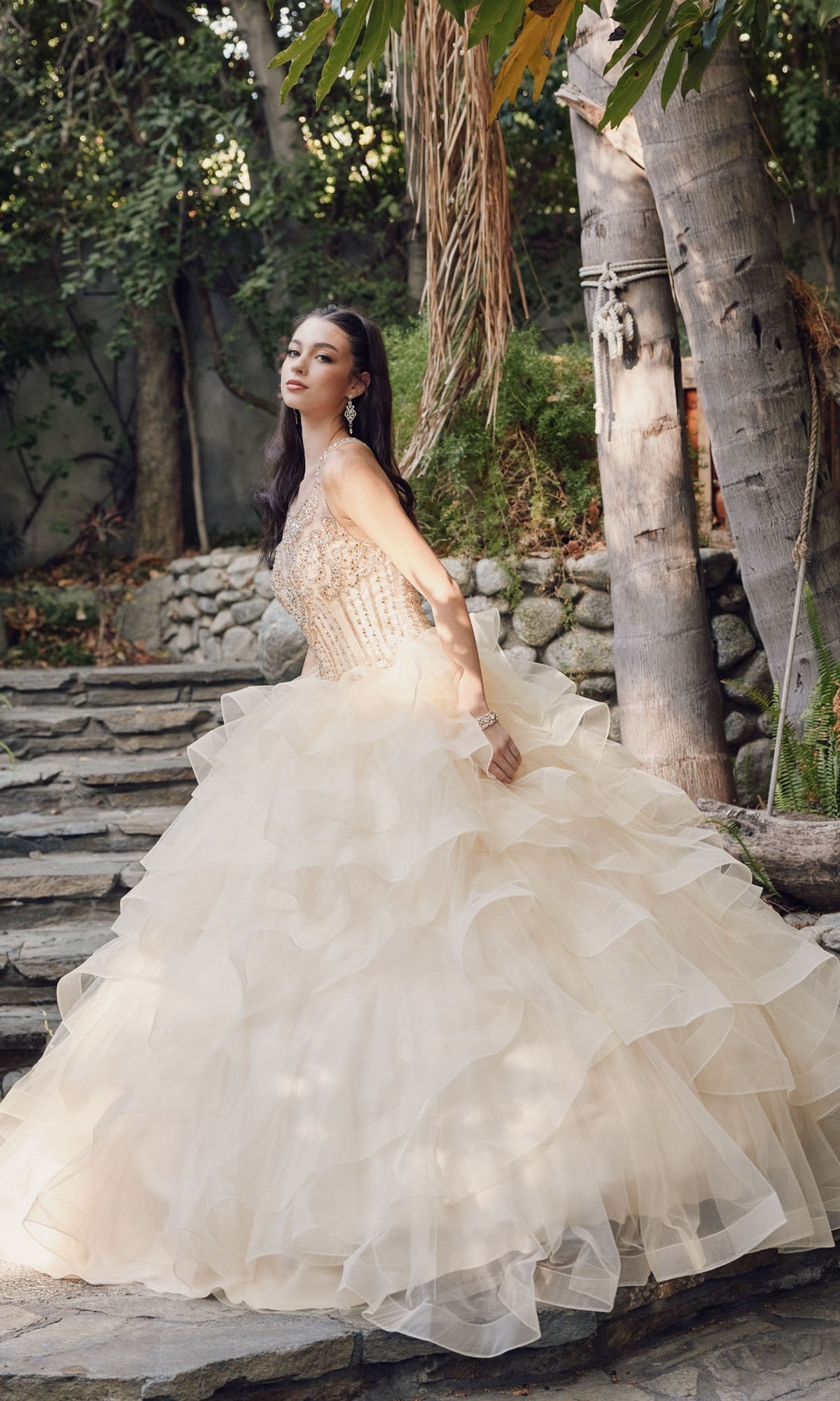 A woman wearing the Quinceanera Dress By Juliet 1423 stands on stone steps outdoors, surrounded by trees and greenery.