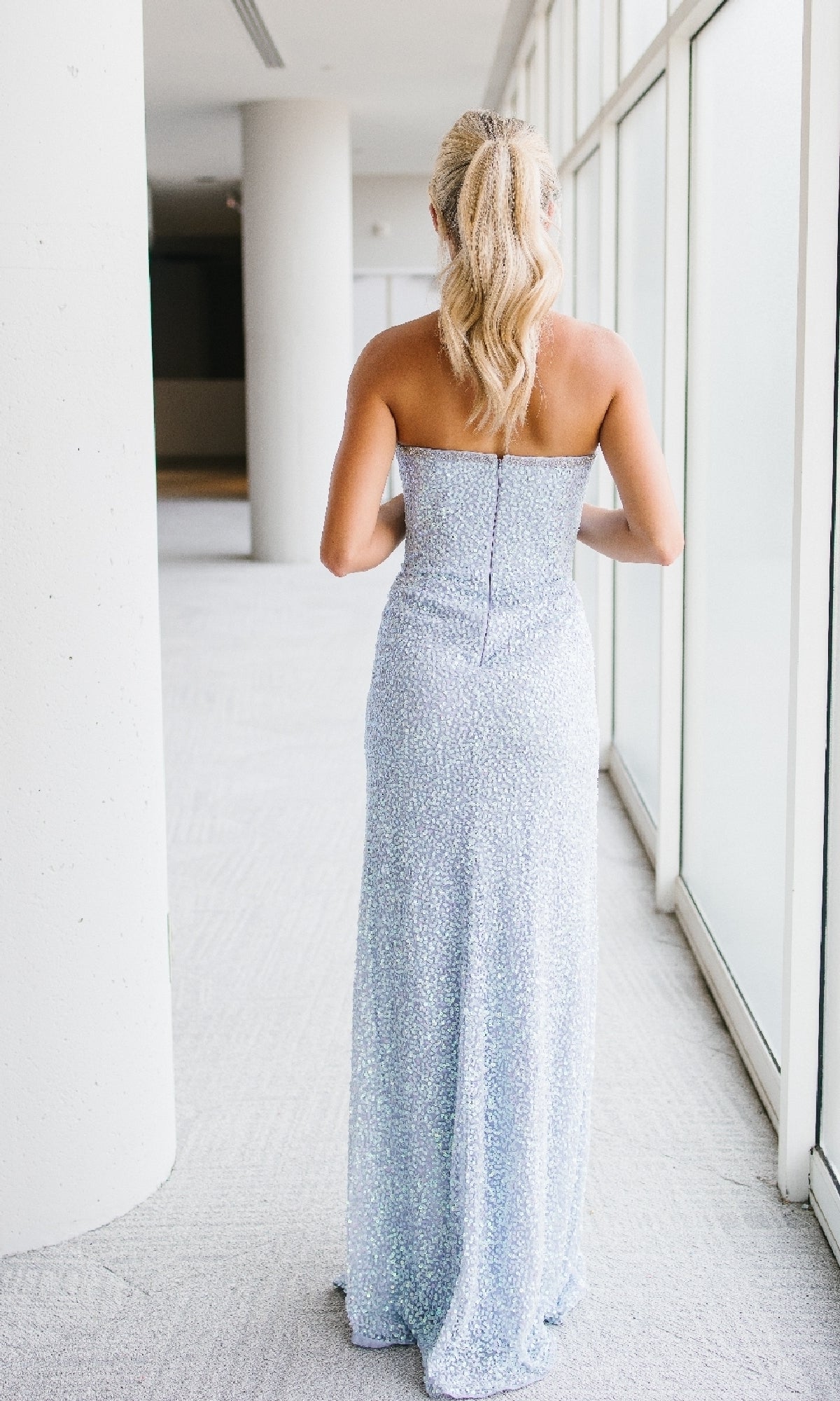 A woman with blonde hair in a ponytail wears the strapless, light blue Long Prom Dress: Aleta 1426 while standing indoors facing large windows.