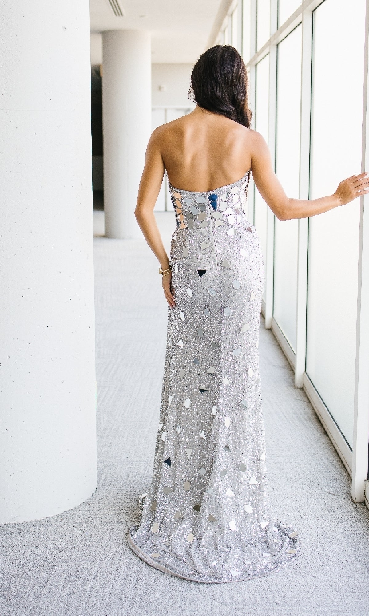 A woman in the Aleta 1429 Long Prom Dress, a reflective silver gown with a sweetheart neckline, stands with her back to the camera, one hand resting on a window ledge in a bright hallway.