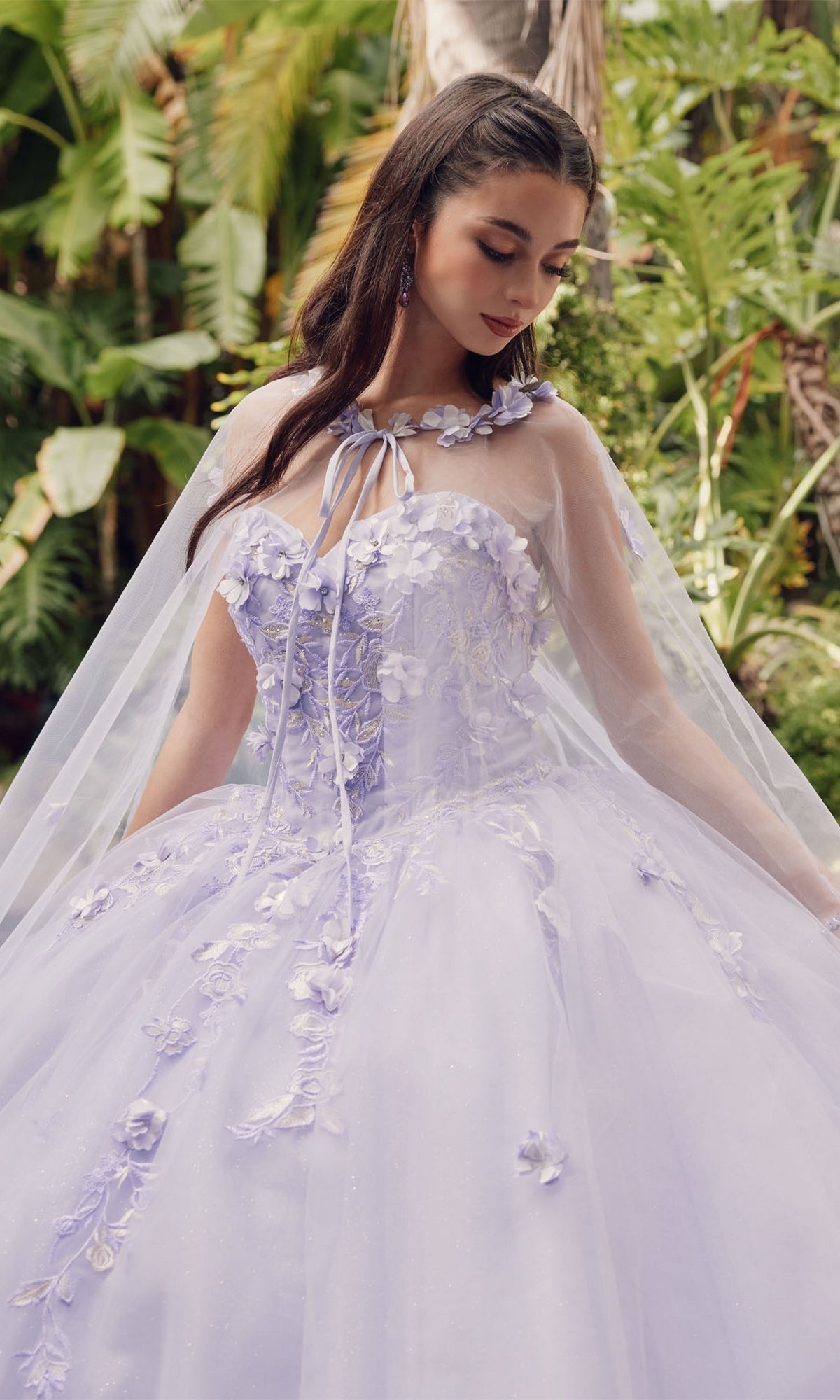 A young woman stands outdoors among green foliage wearing the Juliet 1435 Strapless Ball Gown Quinceañera Dress in lavender, featuring floral appliqués and a sheer cape.