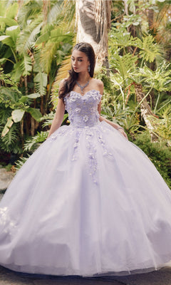 A woman stands outdoors in front of lush green plants and palm trees, wearing the Juliet 1435 Strapless Ball Gown Quinceañera Dress in lavender with floral appliqués.