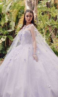 A woman wearing the Juliet 1435 Strapless Ball Gown Quinceañera Dress with floral details and a sheer cape stands outdoors among lush green tropical plants.