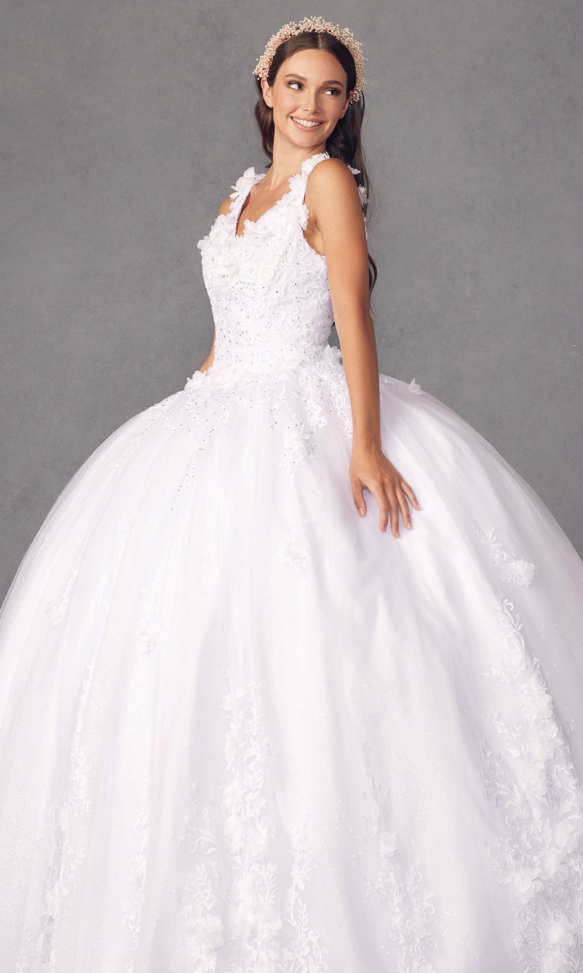 A young woman wearing the Juliet 1437W Traditional White Quinceañera Dress and a decorative headband stands against a gray background, smiling and gazing to the side.