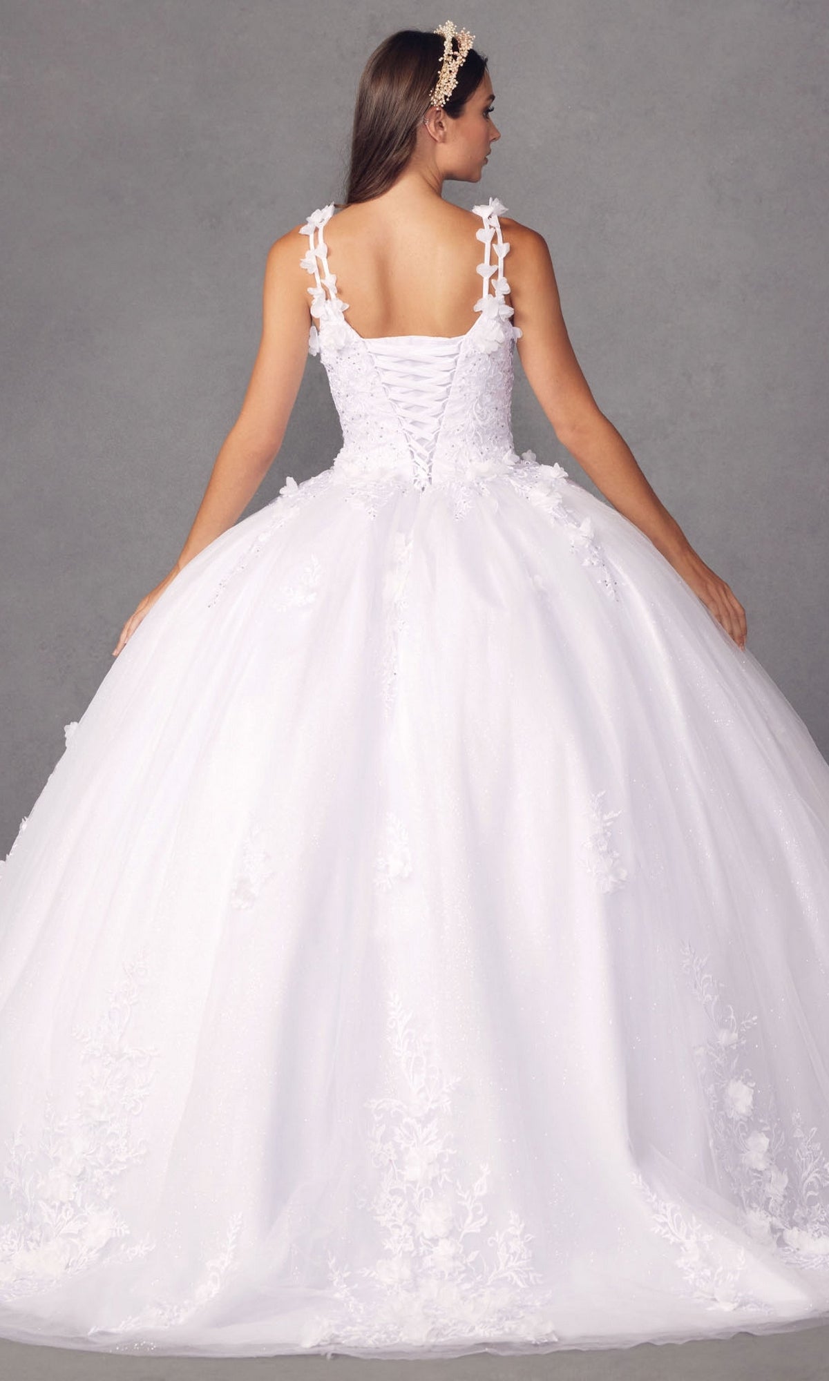 Wearing the Juliet 1437W Traditional White Quinceañera Dress with a lace-up back, floral details, and ball gown silhouette, a woman stands against a neutral gray background facing away from the camera.
