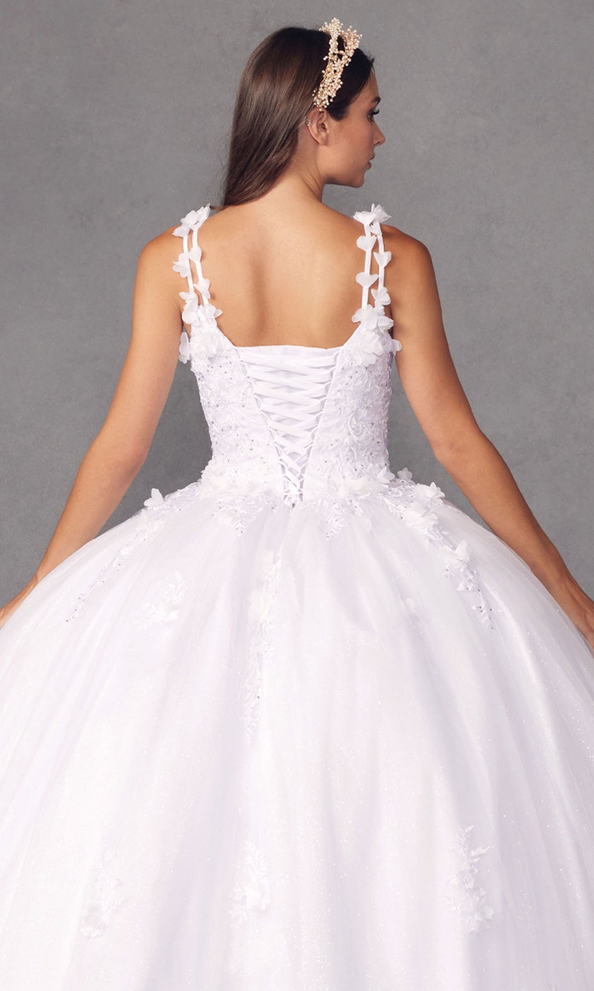 Woman wearing the Juliet 1437W Traditional White Quinceañera Dress, featuring floral appliqué and a lace-up back, stands against a gray background, facing away from the camera.