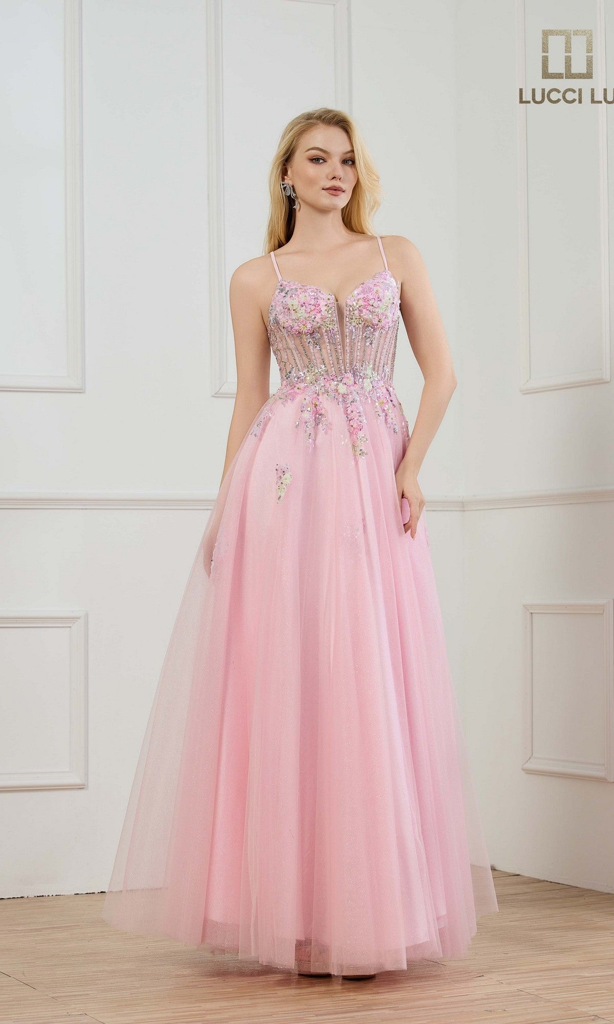 A woman stands in a white room with panel molding, wearing the Lucci Lu 1437—a floor-length, light pink prom dress featuring a fitted sheer corset bodice and delicate embellishments.