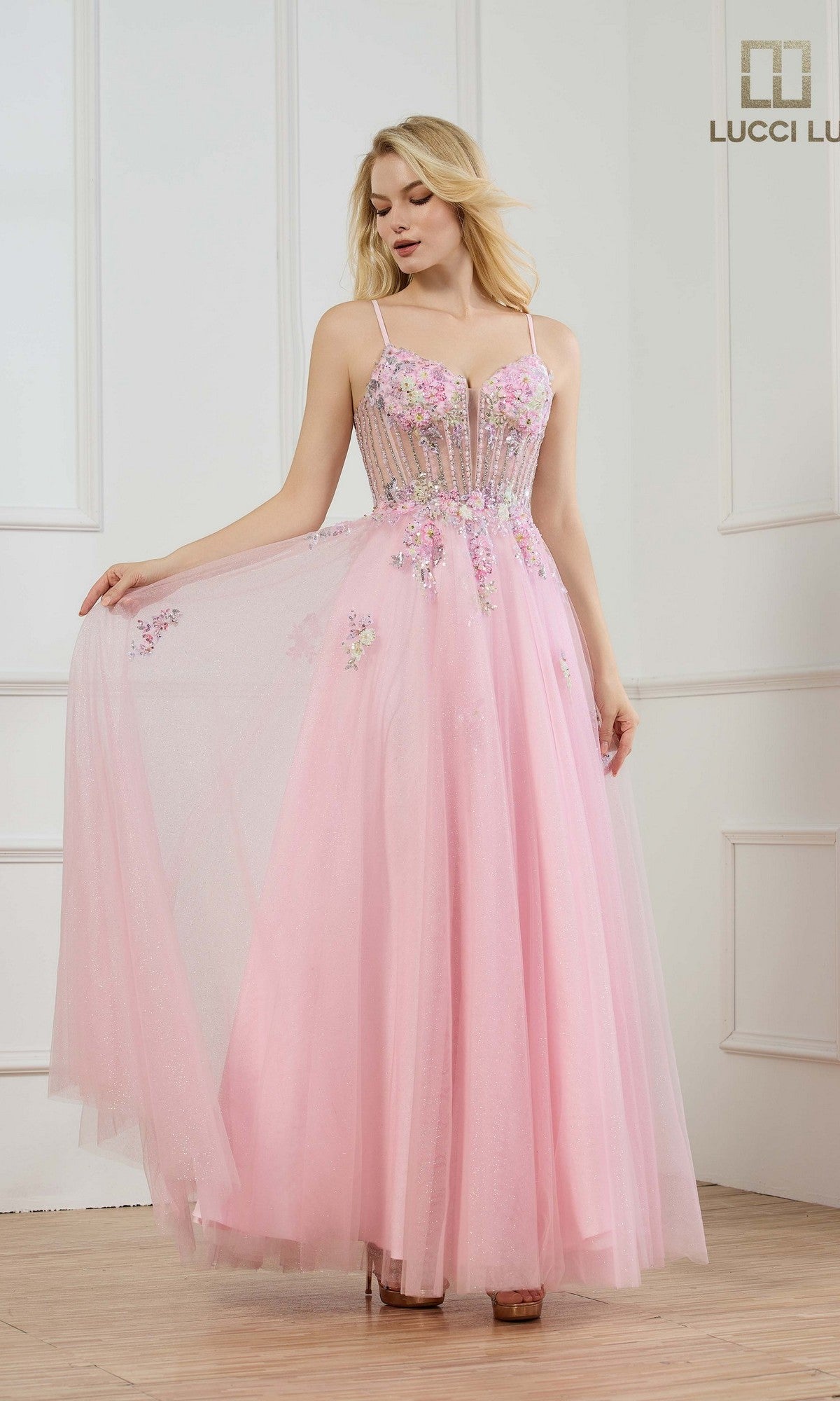 A woman stands in a white-paneled room wearing the Lucci Lu 1437 long pink prom dress, featuring floral embroidery, a sheer bodice, and spaghetti straps.