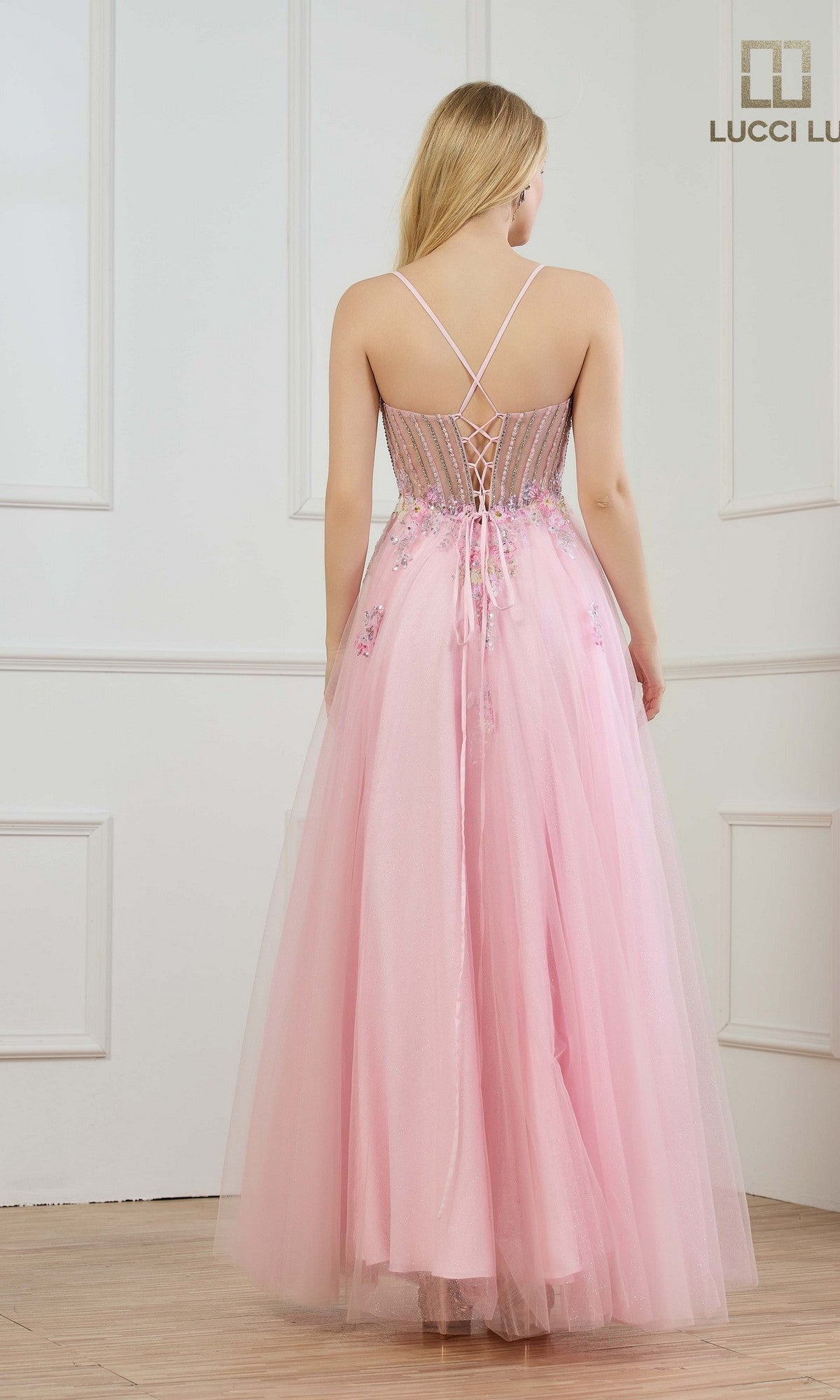 A woman with long blonde hair wears the Lucci Lu 1437, a floor-length light pink prom dress with a sheer corset bodice and thin straps, standing in a white-paneled room.