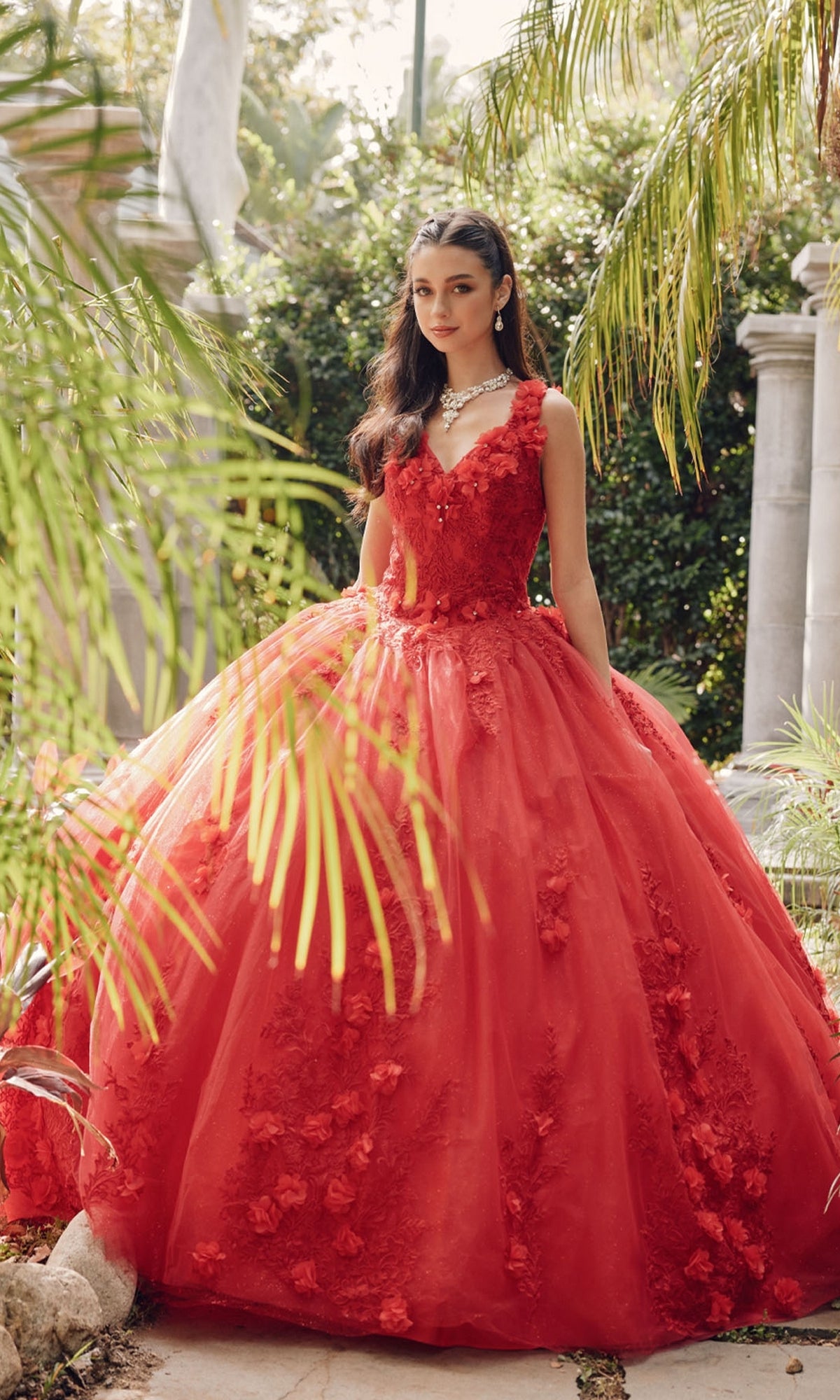 A young woman stands outdoors in the Juliet 1437R Red Ball Gown Quinceañera Dress, featuring floral embellishments and beaded embroidery. She wears a diamond necklace, surrounded by lush greenery.