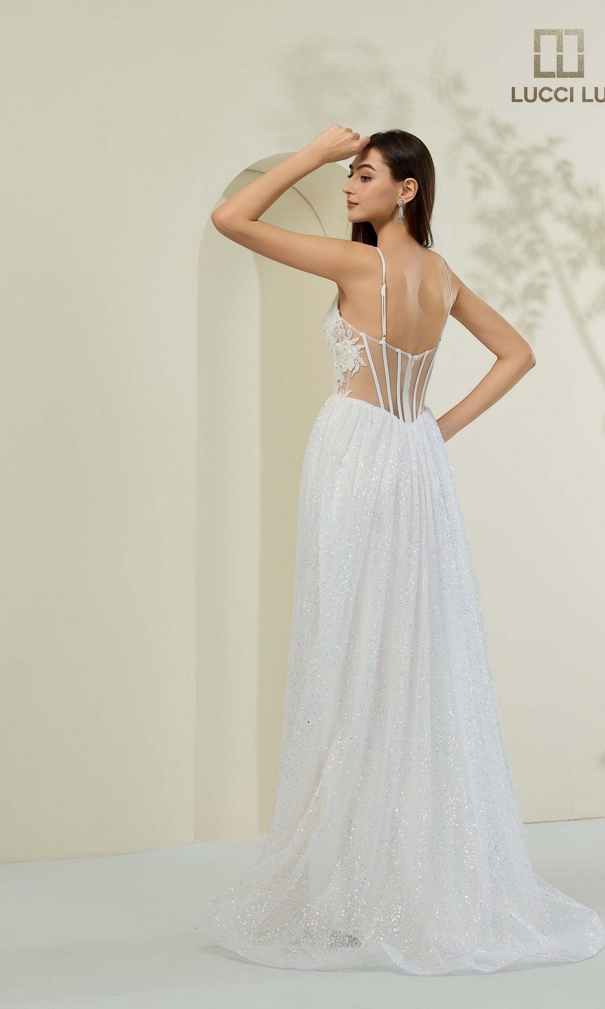 Woman wears the Lucci Lu 1441 long prom dress: a white, floor-length sparkly gown with thin straps, sheer panels, low back, and sleek corset bodice silhouette, standing in a minimalist room with a light background.