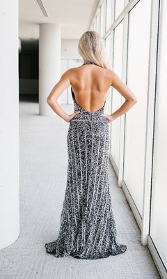 A woman stands with her back to the camera, wearing the Backless Long Sequin Halter Prom Dress: Aleta 1441—a striking floor-length gown—in a brightly lit corridor with large windows.