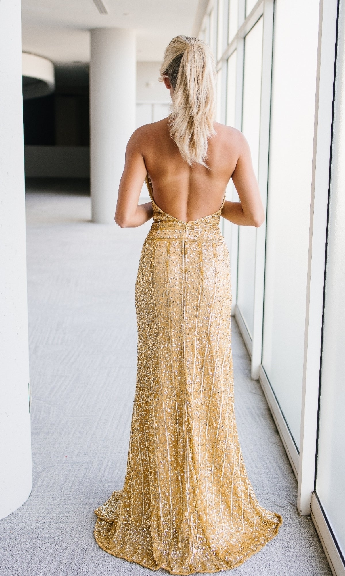 A person with blonde hair in a ponytail wears the Backless Long Sequin Halter Prom Dress: Aleta 1441, standing in a hallway with large windows.