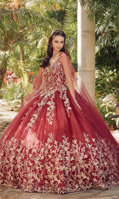 A woman stands outdoors in the Juliet 1442 Quinceañera Dress with Sheer Cape, a voluminous red ball gown featuring intricate white floral embroidery, surrounded by lush greenery.