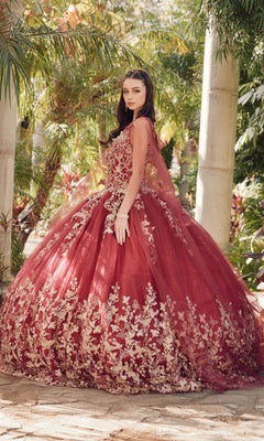 A woman stands outdoors in a garden wearing the Juliet 1442 Quinceañera Dress with Sheer Cape, featuring a voluminous burgundy skirt and gold floral embroidery, looking over her shoulder.
