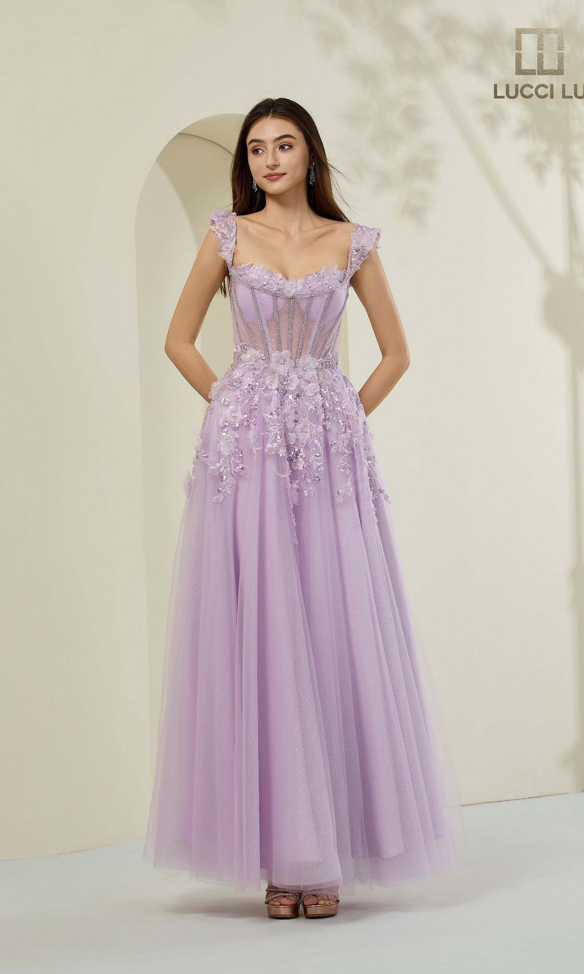 A woman poses in a lilac Lucci Lu 1443 long prom dress featuring embroidered tulle, sheer bodice details, and 3D floral appliques against a neutral background.