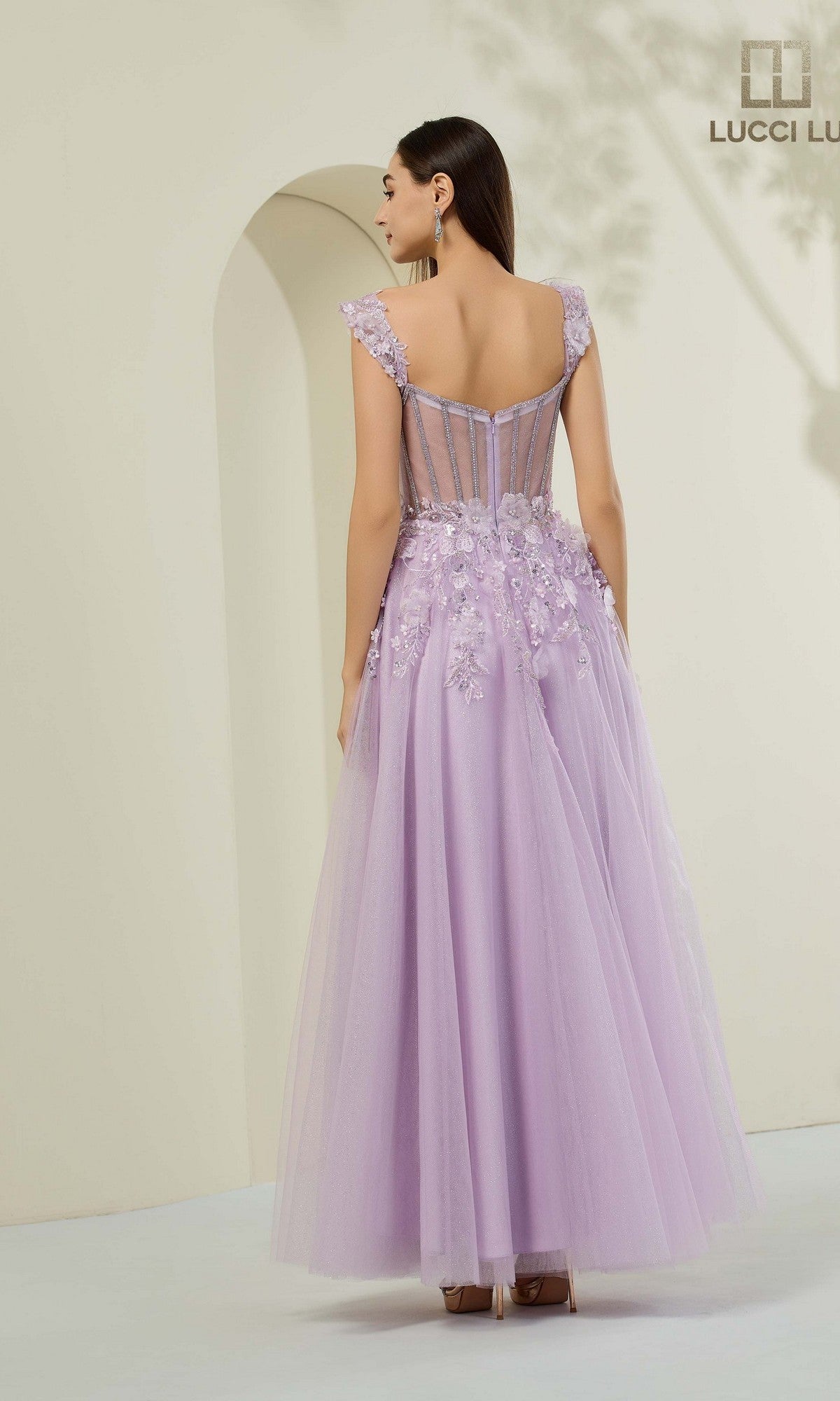 A woman faces away, wearing the Lucci Lu 1443 long prom dress with 3D floral appliques and a sheer back, in a softly lit, neutral setting.