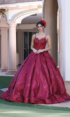 A woman stands outdoors before an arched building in the Quinceanera Dress 1447 by Dancing Queen—a sparkling red lace-embroidered ball gown with thin straps and a matching red flower in her hair.