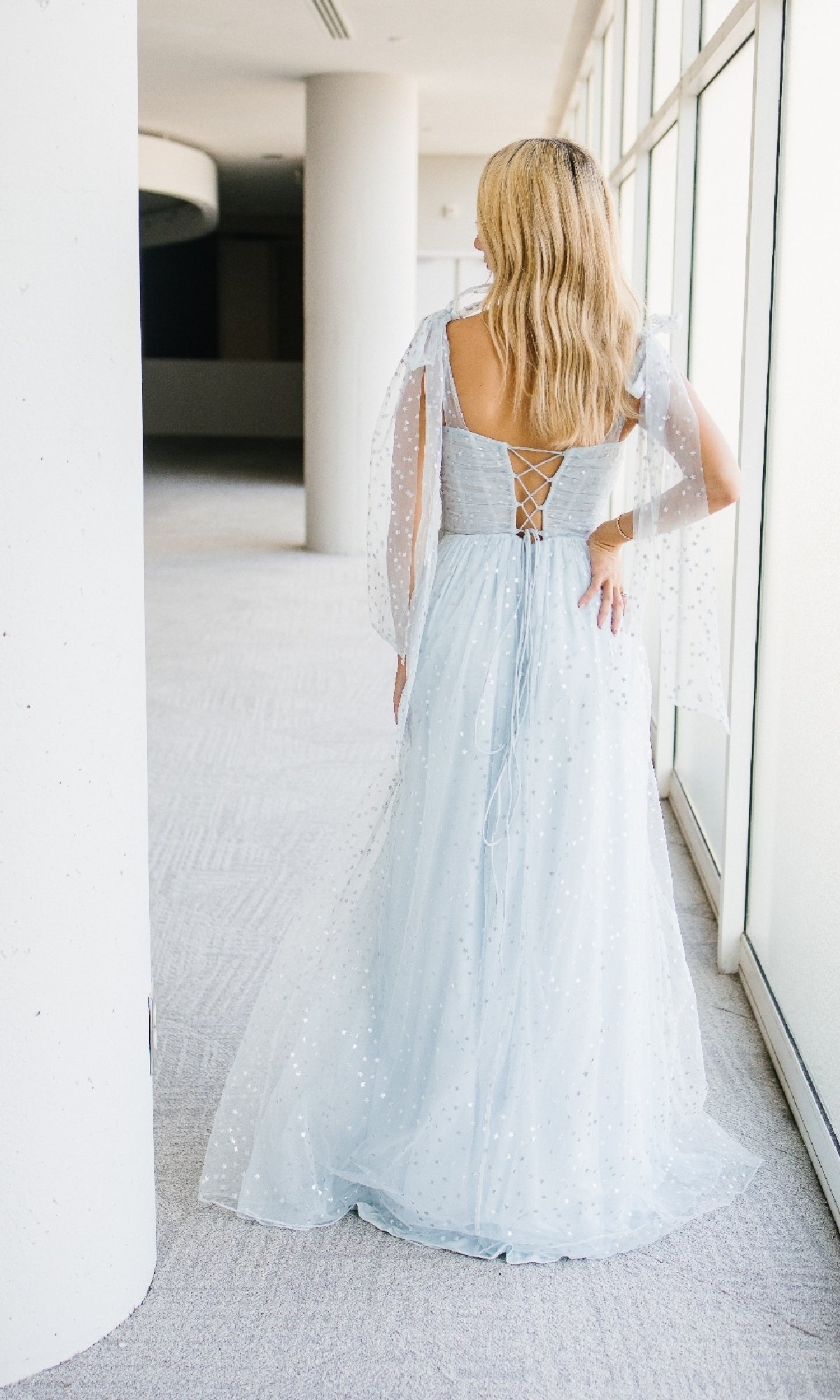 A woman with long blonde hair stands indoors, facing away, wearing the Aleta 1447 Long Prom Dress in light blue, featuring sheer dotted sleeves, a lace-up back, and an elegant A-line silhouette.