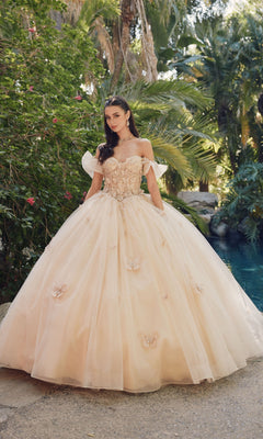 A woman stands outdoors near lush greenery and a pool, wearing the Quinceanera Dress By Juliet JT1448J—a light pink, off-the-shoulder ball gown with a sweetheart neckline and butterfly details.