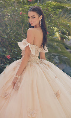 A woman in the Quinceanera Dress By Juliet JT1448J, an off-shoulder a-line ball gown with butterfly details, stands outdoors in light pink, gracefully looking over her shoulder.