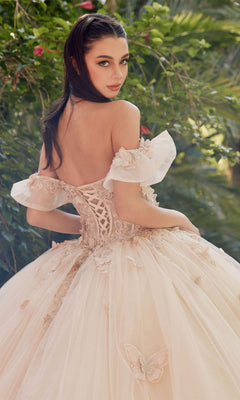 Wearing the Quinceanera Dress By Juliet JT1448J, a woman in an off-the-shoulder a-line ball gown with floral details stands outdoors, glancing over her shoulder, surrounded by lush greenery.