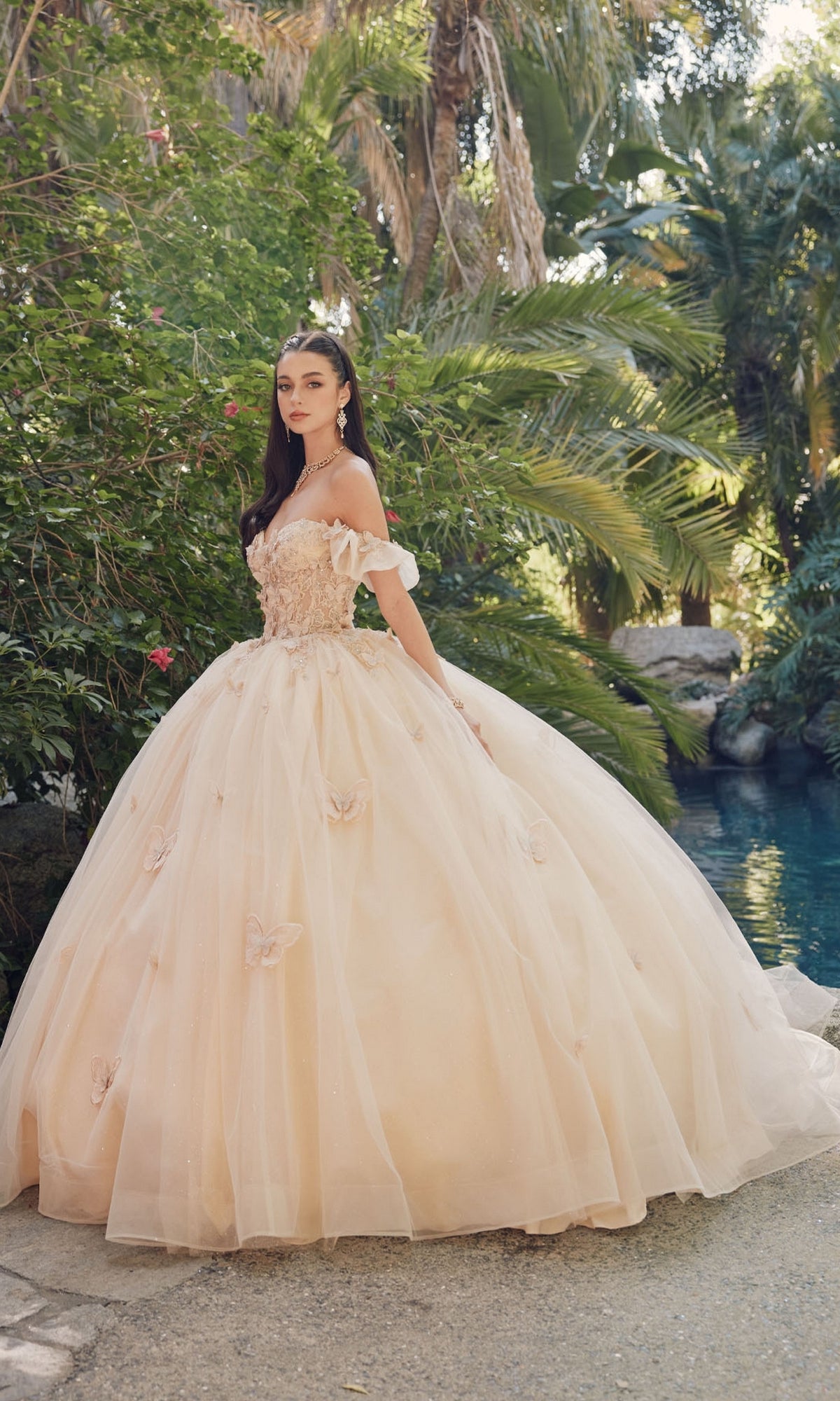 Wearing the Quinceanera Dress By Juliet JT1448J, a woman stands outdoors by greenery and a pool in an A-line ball gown featuring floral details and a sweetheart neckline, gazing at the camera.
