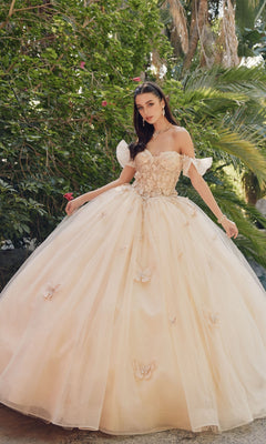 A woman stands outdoors in the Quinceanera Dress By Juliet JT1448J—an off-the-shoulder A-line ball gown with floral and butterfly accents—surrounded by lush green foliage.