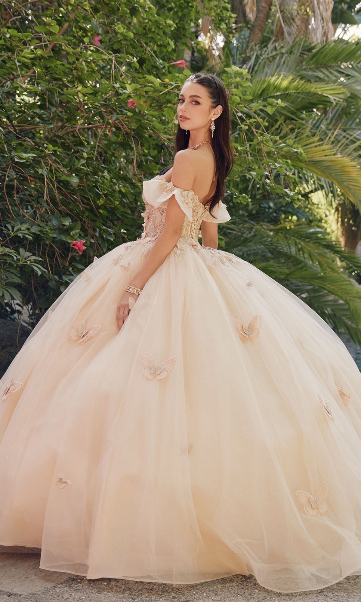 A woman stands outdoors in the Quinceanera Dress By Juliet JT1448J, a light pink off-the-shoulder ball gown with butterfly accents and a full tulle skirt, surrounded by lush greenery.