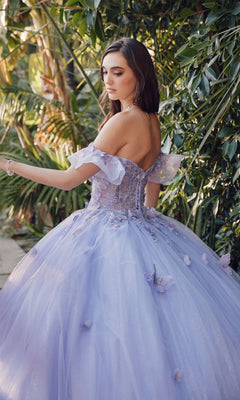 A woman wearing the Quinceanera Dress By Juliet JT1448J, an off-the-shoulder lavender ball gown with a sweetheart neckline, stands outdoors near green foliage, glancing over her shoulder.