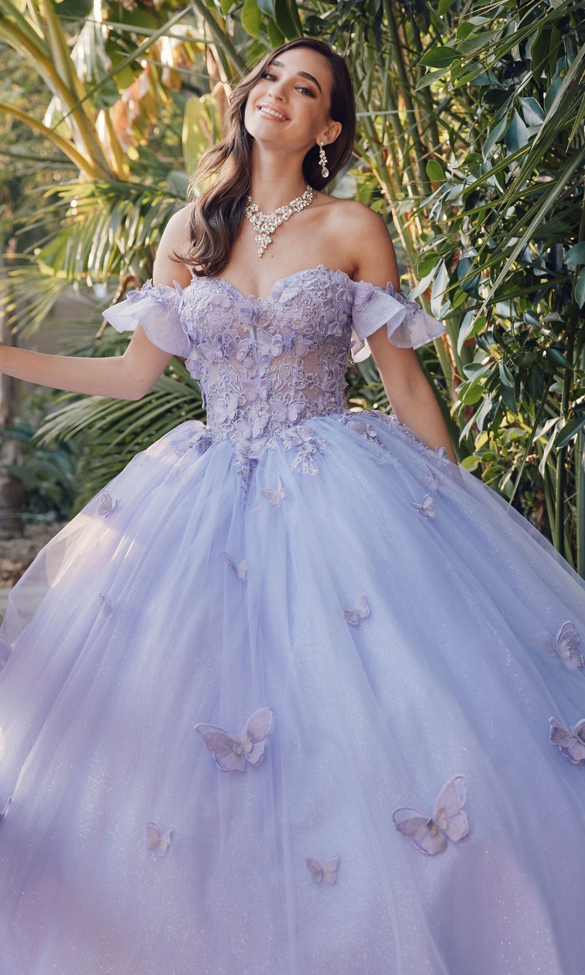 A woman in the Quinceanera Dress By Juliet JT1448J—an off-shoulder lavender ball gown with floral appliqués and butterflies—smiles at the camera while standing among lush green plants.