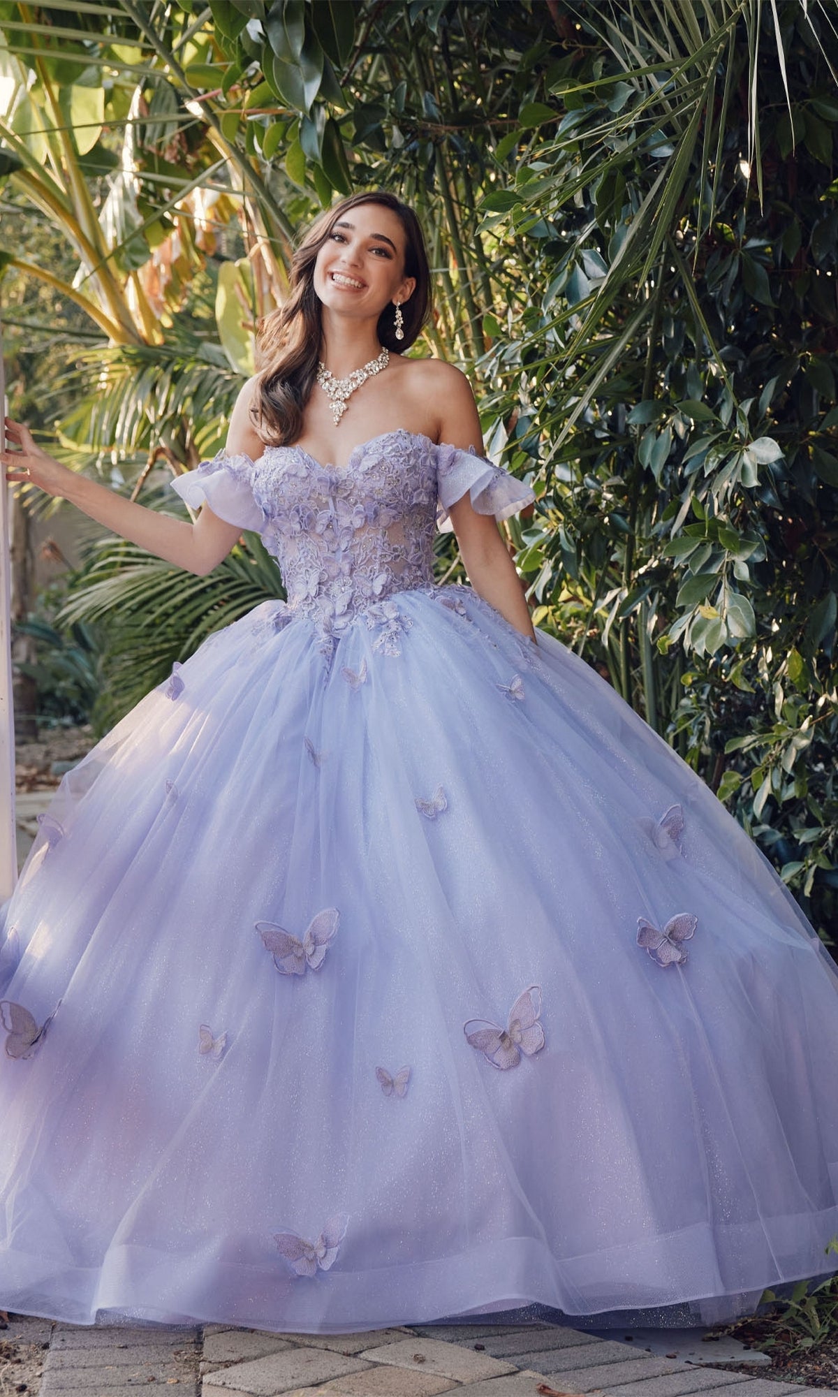 A woman smiles at the camera outdoors, wearing the Quinceanera Dress By Juliet JT1448J—a light purple a-line ball gown with butterfly accents and a sweetheart neckline—surrounded by lush green foliage.
