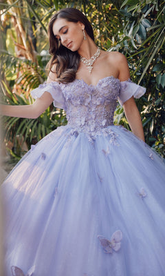 A woman stands outdoors among green plants wearing the Quinceanera Dress By Juliet JT1448J, a lavender off-the-shoulder ballgown with floral and butterfly details, paired with a necklace and earrings.