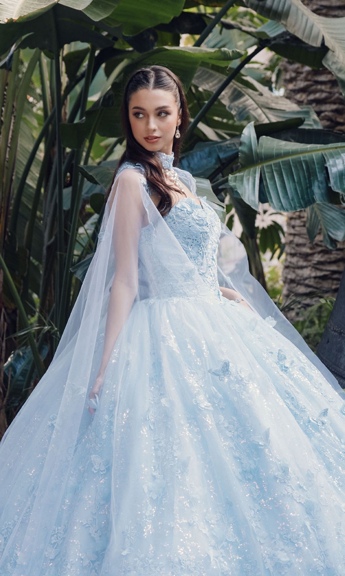 A woman poses outdoors among lush tropical plants in the Quinceanera Dress JT1449J by Juliet, a Bahama blue a-line ball gown with floral embroidery and a sheer cape.