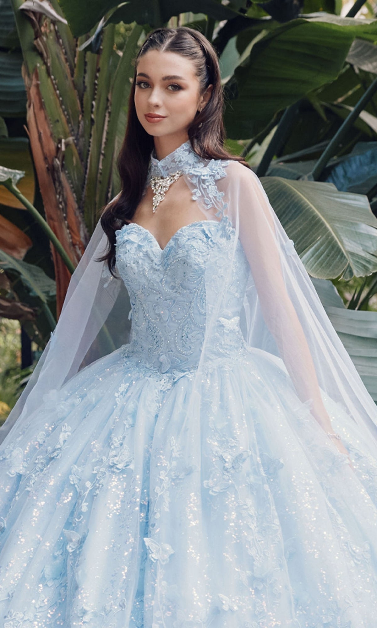 A woman wearing the Quinceanera Dress JT1449J by Juliet in Bahama blue, featuring a strapless sweetheart neckline and sheer cape, stands gracefully before large green leafy plants.