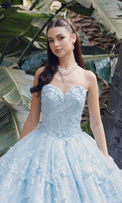 A young woman poses outdoors in front of lush green leaves, wearing the Quinceanera Dress JT1449J by Juliet, a strapless Bahama blue gown with a sweetheart neckline and floral details, accessorized with pearl jewelry.