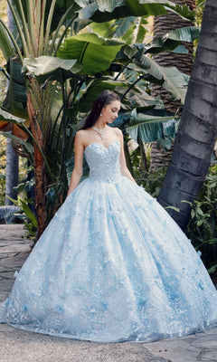 A woman wears the Quinceanera Dress JT1449J by Juliet in Bahama blue, featuring a strapless sweetheart neckline and floral details, as she stands on a stone path among tropical plants.