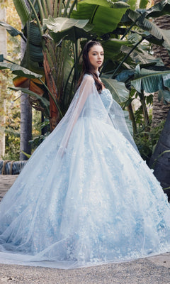 A woman stands outdoors before lush green plants, wearing the Quinceanera Dress JT1449J by Juliet, featuring Bahama blue color, floral details, an a-line ball gown silhouette, and a sheer cape.