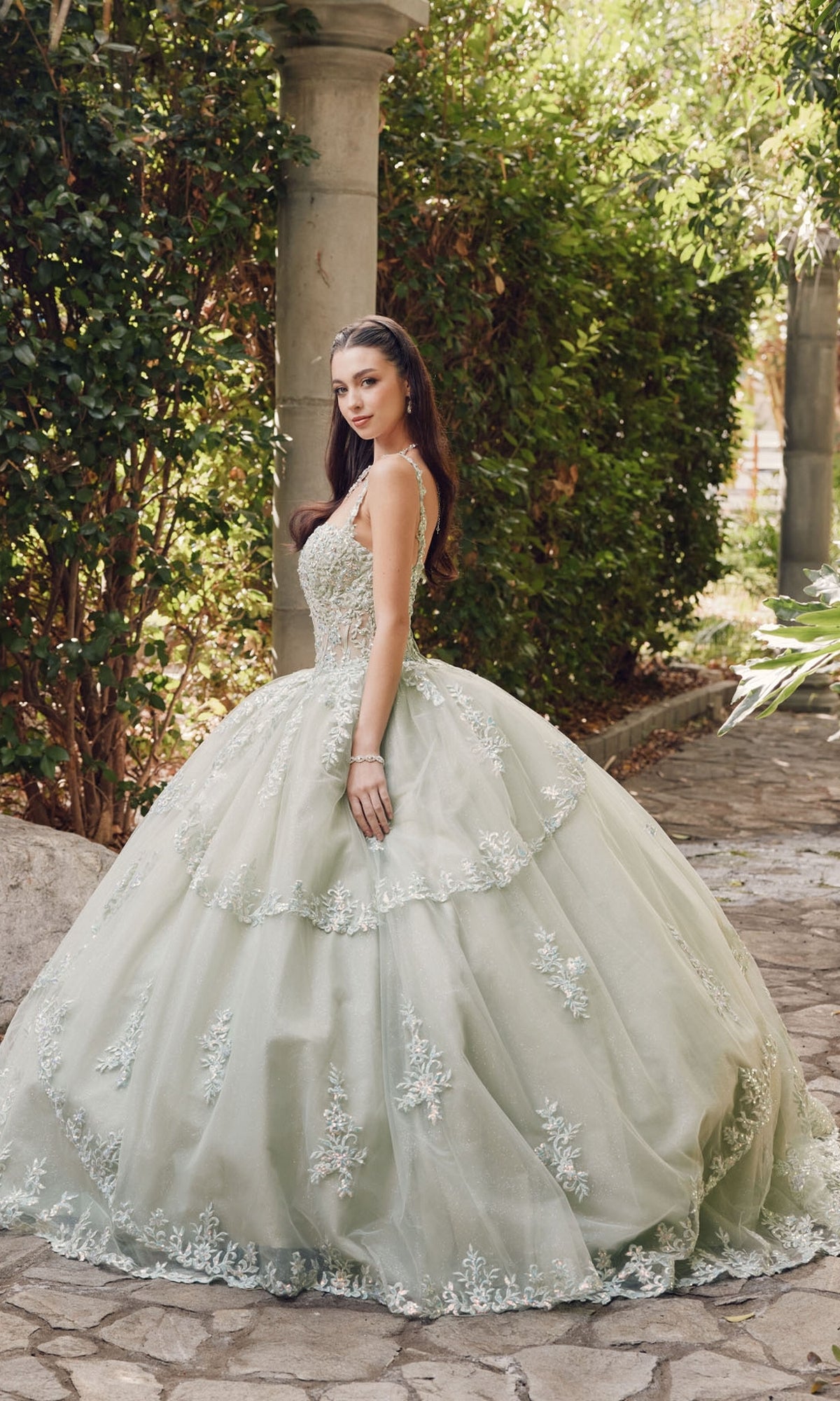 A young woman stands outdoors among greenery and stone columns, wearing the Quinceanera Dress By Juliet JT1450J—a pale green ball gown with a sweetheart neckline, floral appliqués, and a full layered skirt.