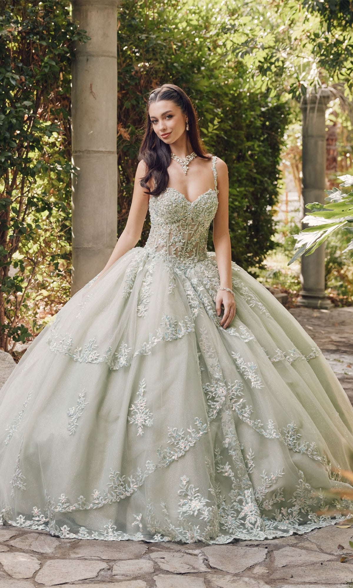 A woman wearing the Quinceanera Dress By Juliet JT1450J, an ornate light green ball gown with a sweetheart neckline and floral embroidery, stands outdoors on a stone path surrounded by greenery.
