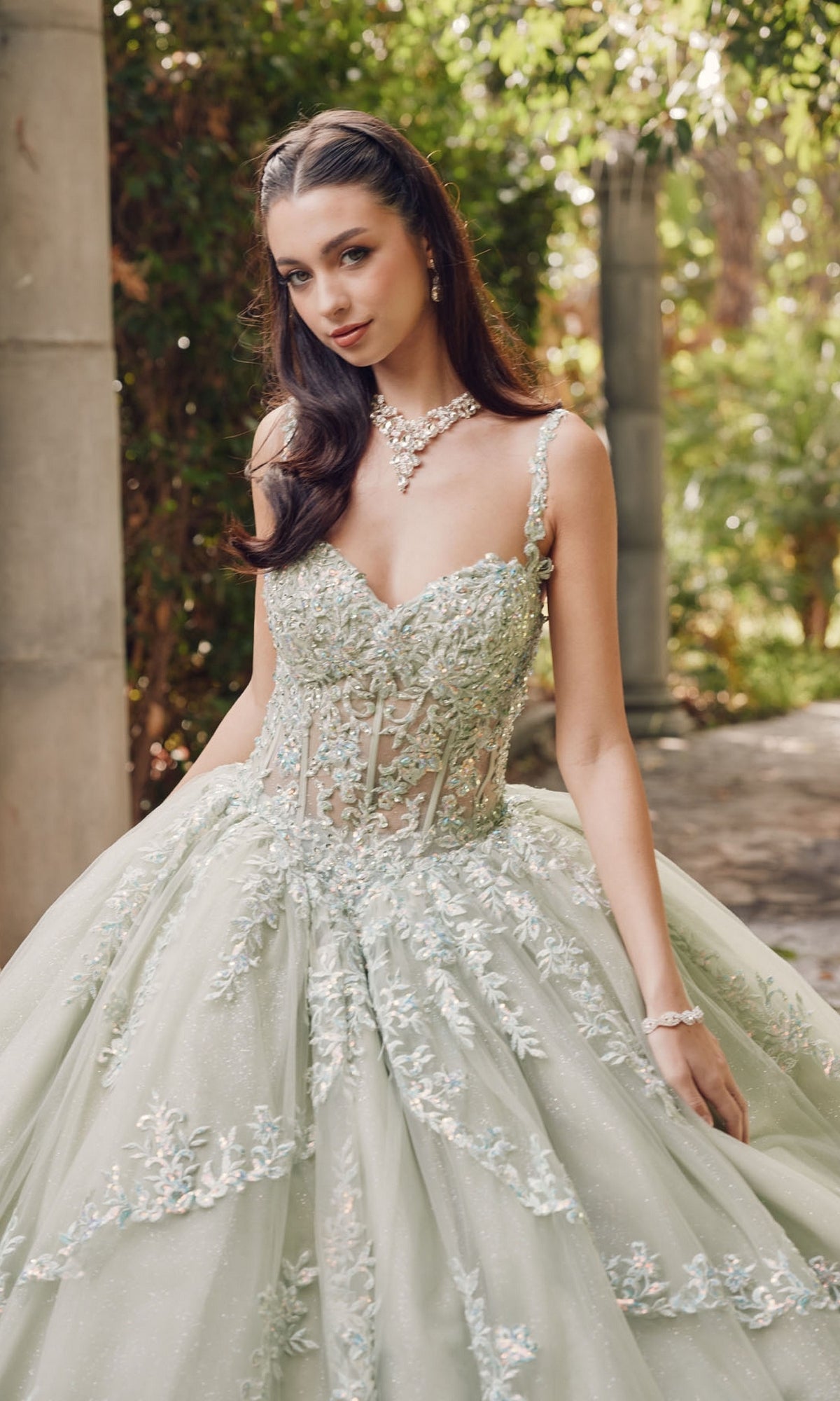 A woman stands outdoors in the Quinceanera Dress By Juliet JT1450J, a light green ball gown with a sweetheart neckline and floral-embroidered tulle, accessorized with a necklace and bracelet.