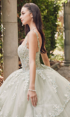 A woman stands outdoors near stone columns and greenery, wearing the Quinceanera Dress By Juliet JT1450J—a pastel ball gown with intricate beading, floral details, and a sweetheart neckline.