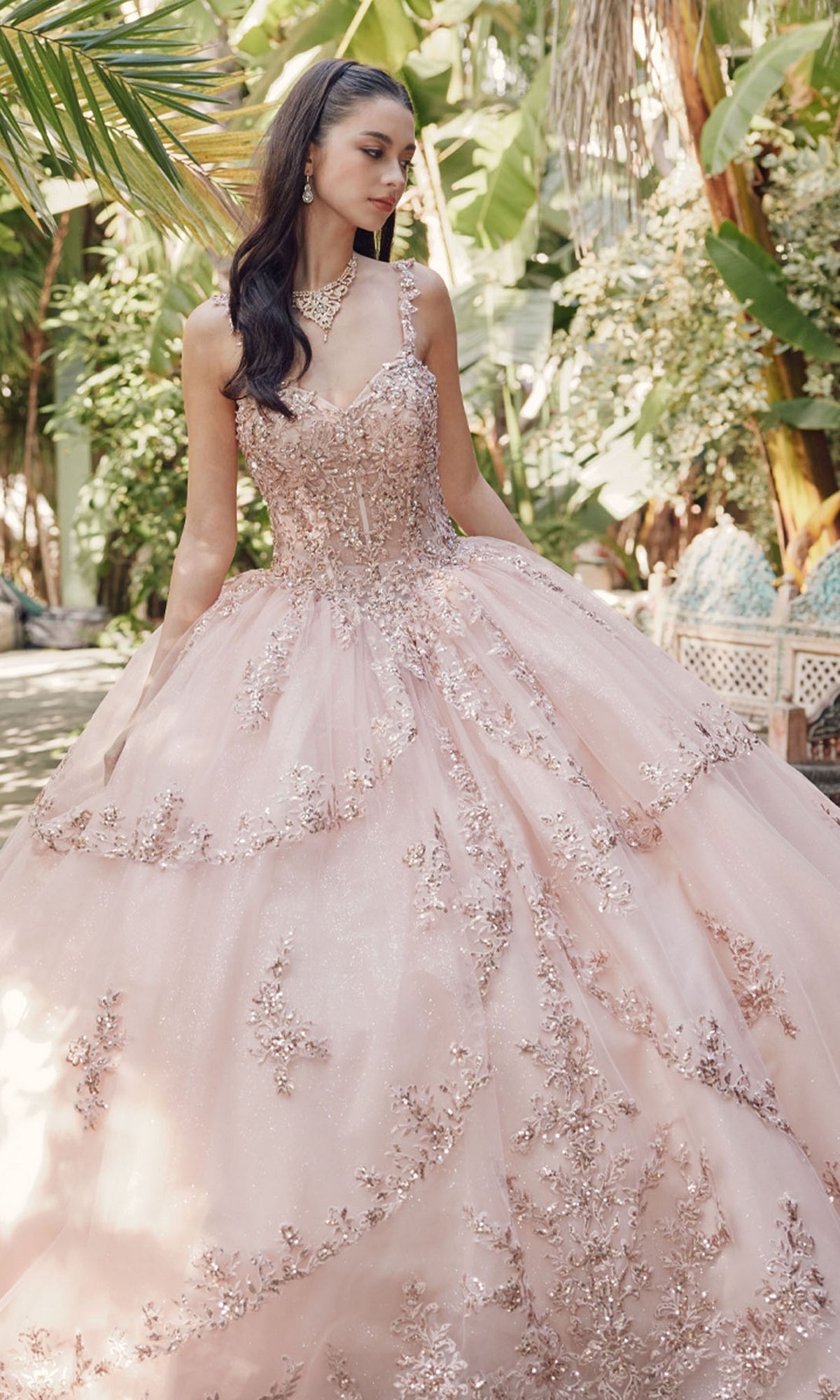 A woman stands outdoors in lush greenery wearing the Quinceanera Dress By Juliet JT1450J, a pale pink ball gown with a sweetheart neckline and floral-embroidered tulle, accessorized with jewelry.