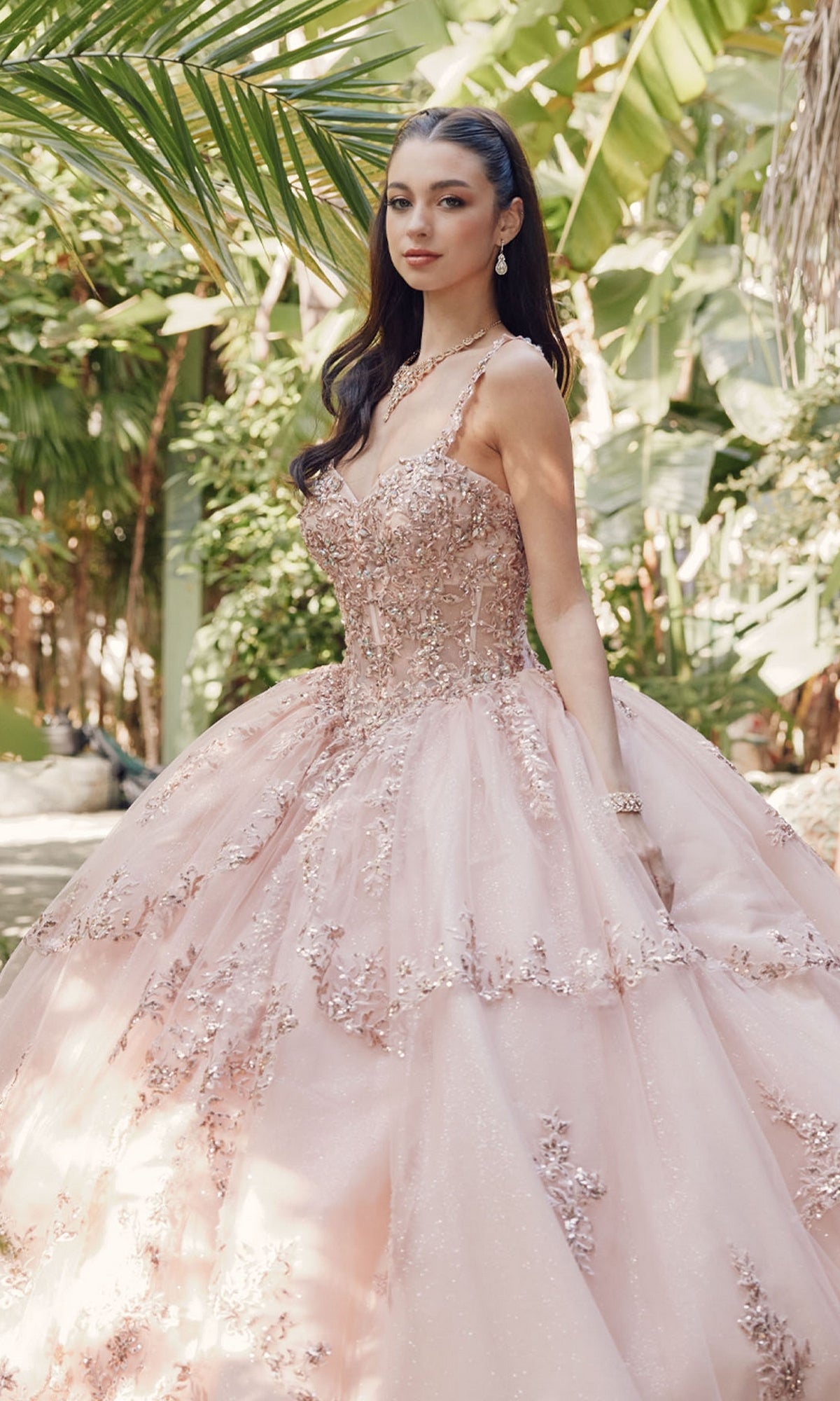 A woman stands outdoors in the Quinceanera Dress By Juliet JT1450J, a light pink ball gown with a sweetheart neckline and floral embellishments, surrounded by lush greenery and elegant jewelry.